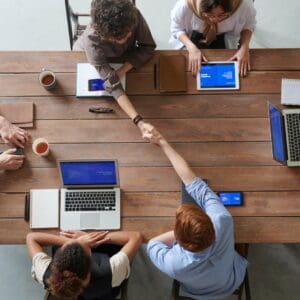 Six people sitting around a table, working on laptops and tablets, and two of them are shaking hands across the table.