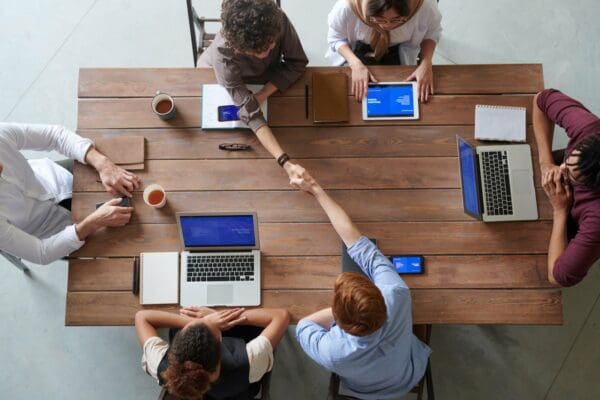 Six people sitting around a table, working on laptops and tablets, and two of them are shaking hands across the table.