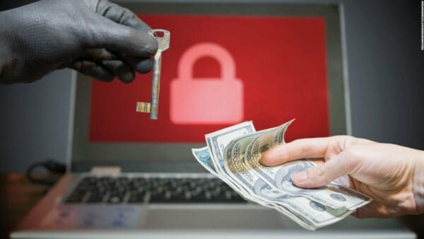 A locked computer on a desk, with a hacker exchanging the key to the computer for money from the owner