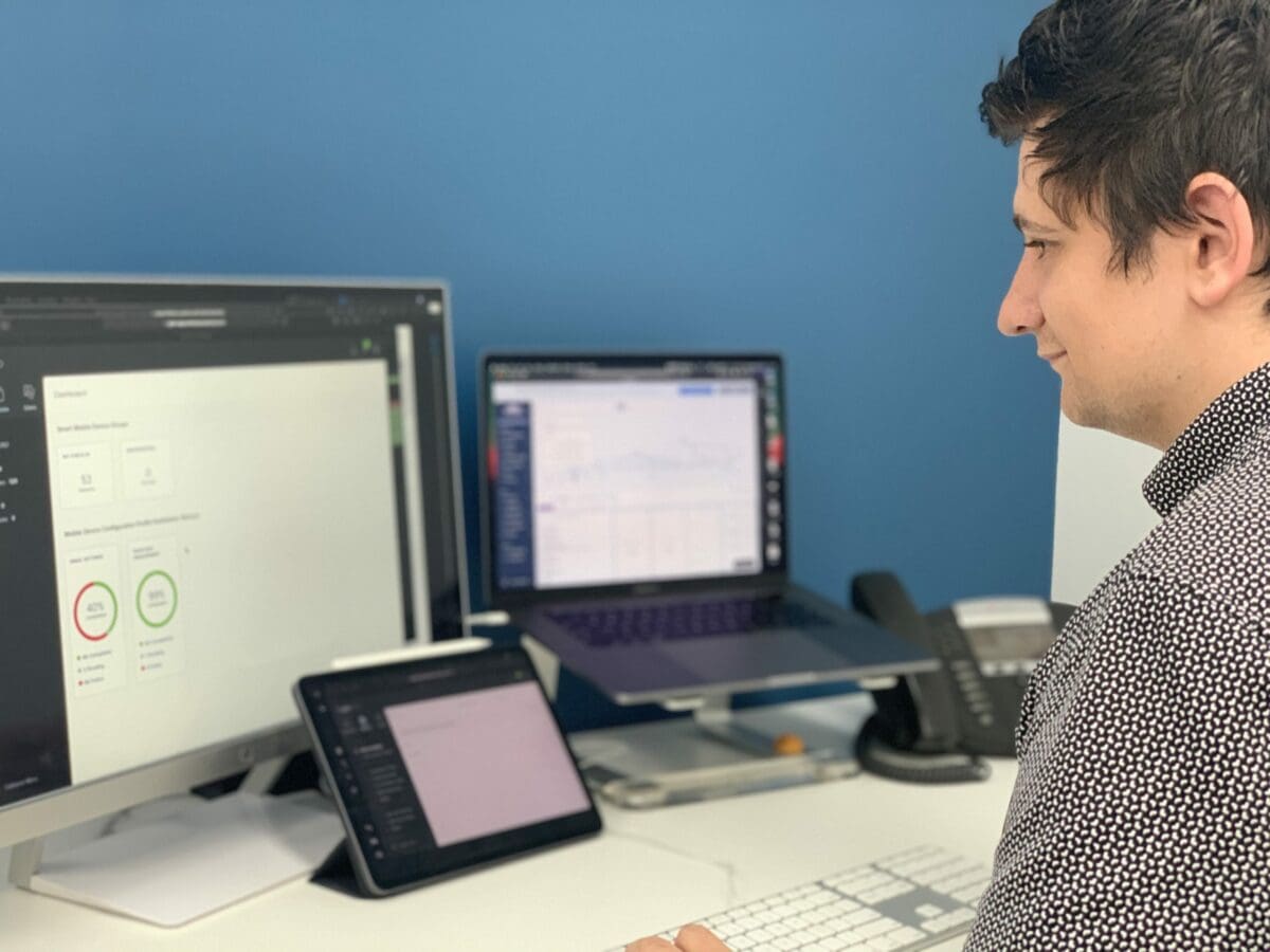 A SupportMyMac employee sitting at his desk at work, looking at two computer screens and an iPad, monitoring the Mobile Device Management (MDM) solution