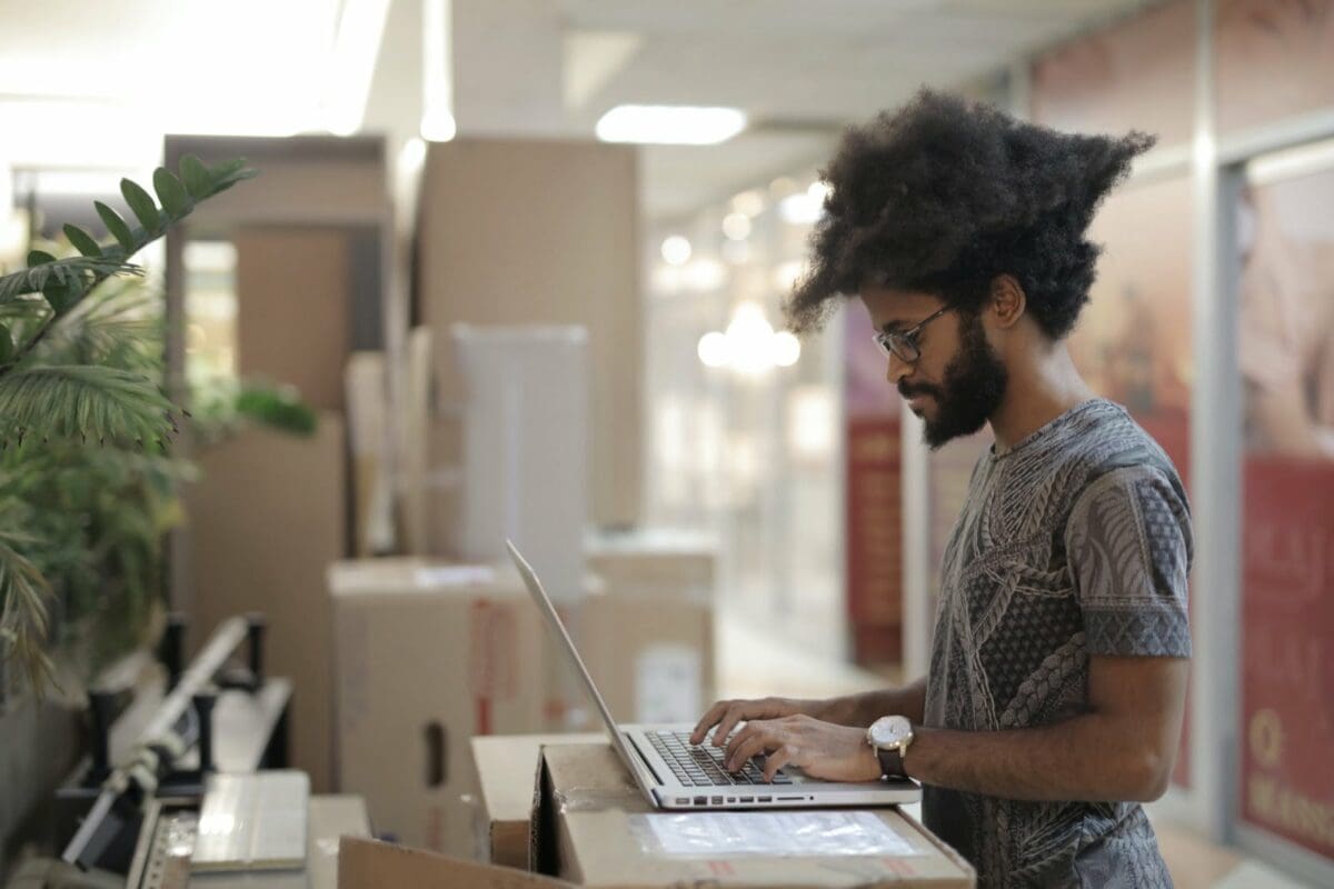 Focused black man working on a laptop in an office space, surrounded by cardboard boxes