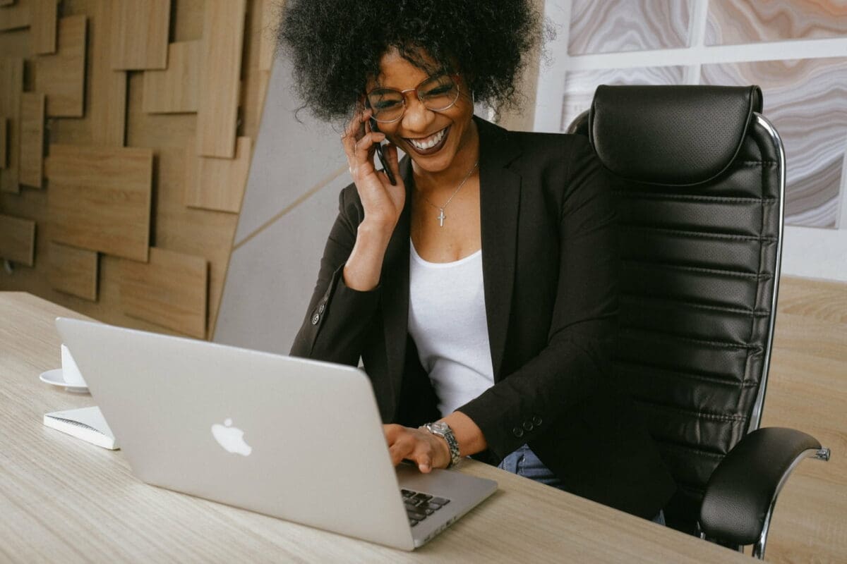 Smiling, ethnic woman in a black blazer sitting on black office chair looking at her Apple computer while talking on her iPhone