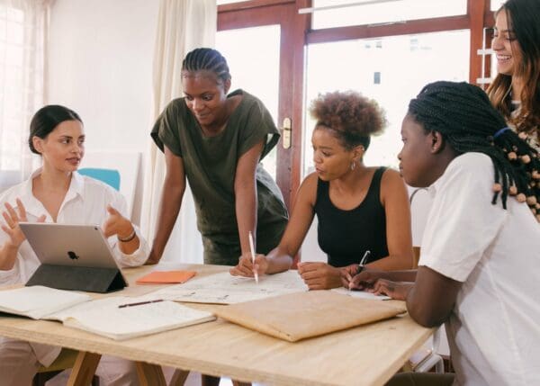 Five women of varying ethnicities sitting and standing around a big boardroom table of what looks like an architect or art firm, looking at an iPad mobile device
