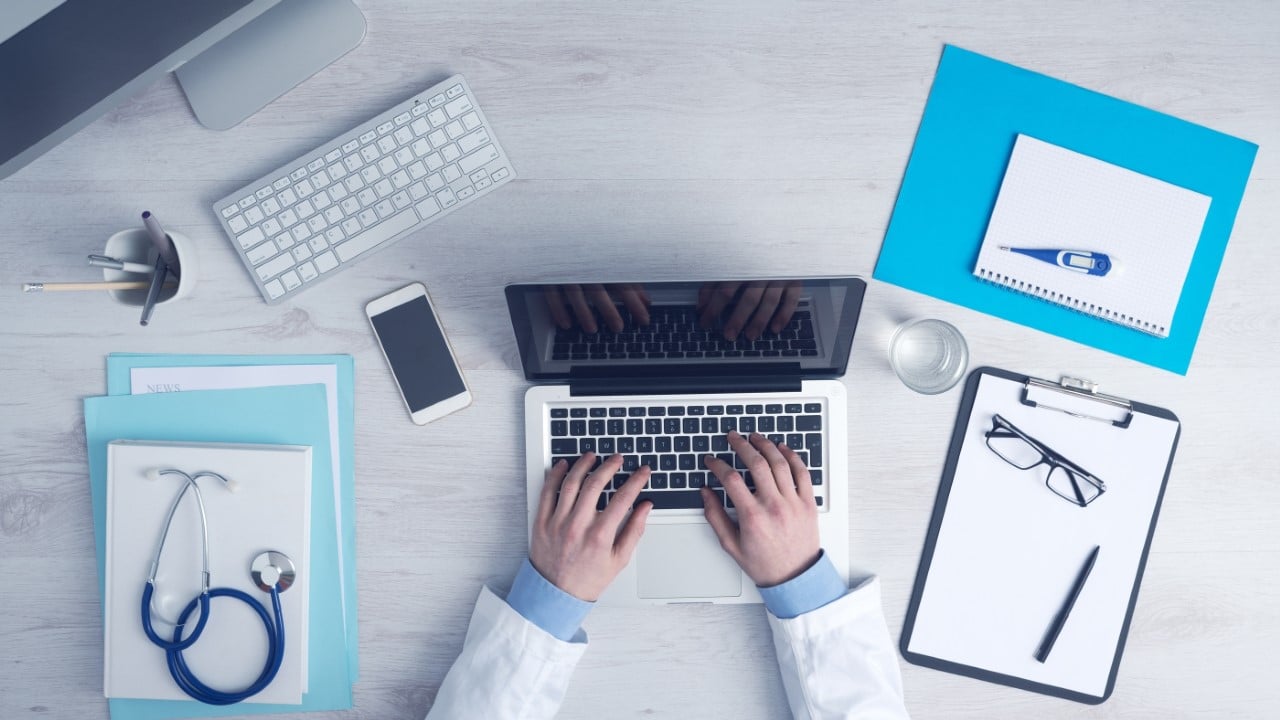 Bird's eye view of a doctor's arms typing on an Apple laptop, on a desk surrounded by patient charts, a stethoscope, a clipboard, glasses and a thermometer