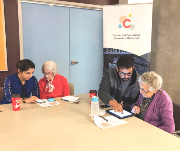 Two Connected Canadians volunteers sitting at a table with two seniors, helping them use tablets.