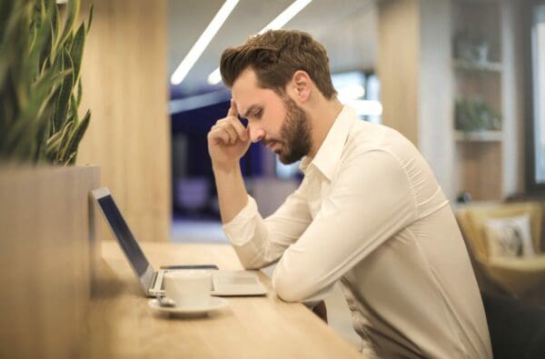 Bearded man in a white collared shirt, sitting at his computer with his hand on his temple, looking downwards.